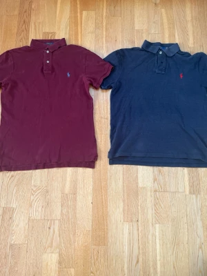 Två pikétröjor från Polo Ralph Lauren - Den ena pike tröjan är S men båda är lika stora. Säljer två klassiska pikétröjor från Polo Ralph Lauren. En är vinröd med blå logga och den andra är mörkblå med röd logga. Båda har krage, knappar framtill och korta ärmar. Perfekta för en stilren och avslappnad look.