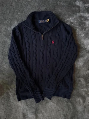 Mörkblå kabelstickad tröja Polo Ralph Lauren - Kabelstickad mörkblå tröja från Polo Ralph Lauren med halv dragkedja och hög krage. Tröjan har det klassiska röda Polo-logot broderat på bröstet och ribbade muddar vid ärmslut och nederkant. Perfekt för en stilren och avslappnad look.
