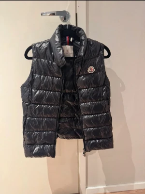 Svart dunväst från Moncler - Snygg svart dunväst från Moncler med glansig finish och klassisk logga på bröstet. Västen har hög krage, dragkedja framtill. 