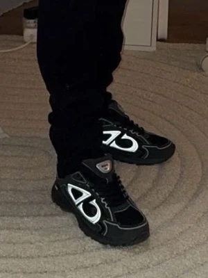 B30 skor - Säljer ett par svarta sneakers från b30 med feta silvriga detaljer på sidorna och svart sula. Skorna har en sportig vibe med snörning och reflekterande partier. Perfekta för dig som vill sticka ut med en unik look.