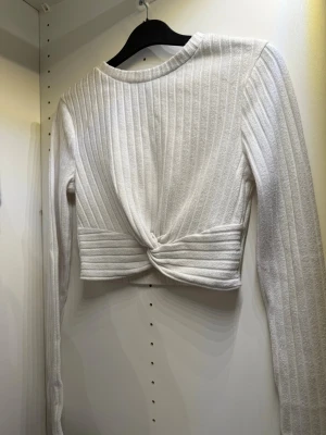 Vit ribbad croppad topp med knut - Säljer en vit croppad topp med lång ärm och snygg ribbad struktur. Toppen har en twistad knutdetalj framtill som ger en cool look. Perfekt till höga jeans eller kjol. Superenkel att styla och passar till det mesta!