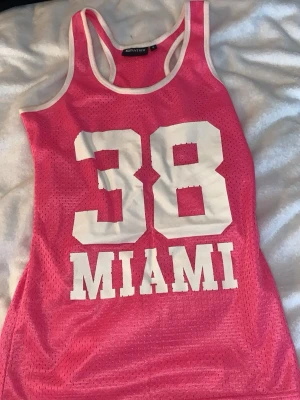 Rosa mesh-linne från Panther - Snyggt rosa mesh-linne från Panther med vita kanter och stora vita tryck på framsidan: siffran 38 och texten MIAMI. Sportig design i luftig polyester som passar perfekt till träning eller streetwear. Ärmlös modell med rund halsringning. Köptes i Belgien 