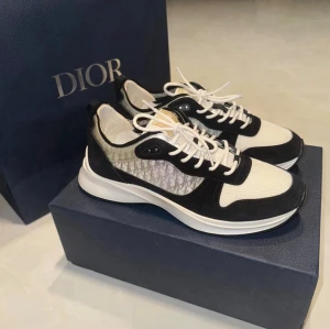Dior B25 Trainers - Snygga Dior sneakers i svart och vitt med klassisk logotyp på sidan. Ovandelen är i mesh och läder, med vita snören och robust sula. Perfekta för dig som vill ha en lyxig och trendig look. Diskreta detaljer och modern design.