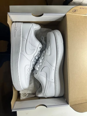 Nike Air Force 1 vita sneakers - Klassiska Nike Air Force 1 sneakers i helvitt läder med perforerad tå, tjock sula och ikoniska swoosh-loggan på sidan. Skorna har vita skosnören och metallbricka med 'AF1' på snörningen. Perfekta för en clean och stilren look.