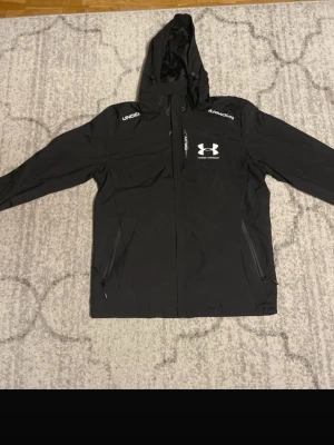Svart vindjacka från Under Armour - Snygg svart vindjacka från Under Armour med huva och dragkedja framtill. Jackan har två fickor med dragkedja och vita logotyper på bröstet och axlarna. Tillverkad i lätt, vattenavvisande material med meshfoder för extra komfort.                                                                 (Nypris: 1500kr❌ vs  Mitt pris: 900kr✅)