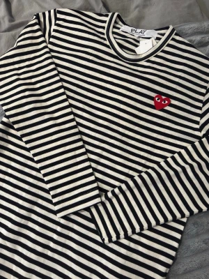 Randig långärmad topp Comme des Garçons - Säljer en svartvit randig långärmad topp från Comme des Garçons Play med det ikoniska röda hjärtat broderat på bröstet. Klassisk passform och rund halsringning. Perfekt för dig som gillar streetstyle och vill ha något tidlöst och trendigt. Tröjan är i storlek L men dem sitter alltid en storlek mindre. Så passar M