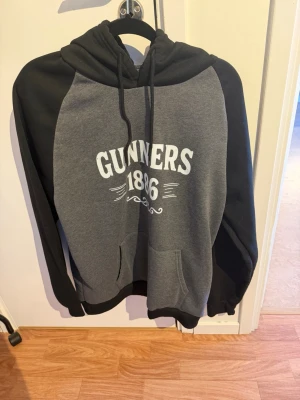 Grå och svart hoodie Gunners 1886 - Snygg hoodie i grått och svart med texten 'Gunners 1886' på bröstet. Tröjan har huva med dragsko och en stor magficka framtill. Perfekt för dig som gillar sportig och avslappnad stil. Materialet känns mjukt och bekvämt.