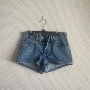 Blå jeansshorts med broderade fickor - Blå jeansshorts från 157 med hög midja och knappar framtill. Baksidan har två bakfickor med mörkblå broderade detaljer.