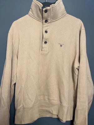 GANT Vintage tröja, 100% bomull – Mycket fint skick - Stilren och klassisk GANT Vintage tröja i storlek M.  Färg: Ljusbrun/beige – varm och lätt att matcha Material: 100% bomull – mjuk och skön kvalitet Design: Hög krage med både knappar och dragkedja undertill, praktisk och snygg detalj  Skick: Mycket fint vintage-skick, välbevarad och utan defekter  Snygg tröja som passar perfekt till höst och vintern.
