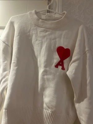 Vit stickad tröja från Ami Paris - Vit stickad tröja från Ami Paris med röd hjärt- och A-detalj på bröstet. Tröjan har en bred ribbad mudd vid hals, ärmslut och nederkant. Oversized passform och mjukt material som känns lyxigt och trendigt.
