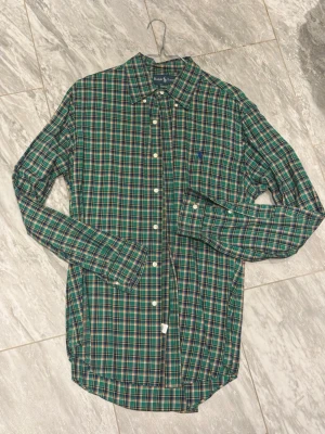 Grön rutig skjorta från Ralph Lauren - Säljer en grön rutig skjorta från Ralph Lauren i modellen Custom Fit. Skjortan har klassisk krage, långa ärmar och knappar framtill. Materialet är mjuk bomull och den har en liten broderad logga på bröstet. Perfekt för dig som gillar stilrena och tidlösa plagg.