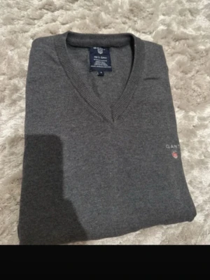 Grå stickad tröja från Gant - Mörkgrå stickad tröja från Gant med V-ringning och diskret logga broderad på bröstet. Tröjan har långa ärmar och är tillverkad i mjukt material som passar perfekt till lager-på-lager. Klassisk och stilren design.