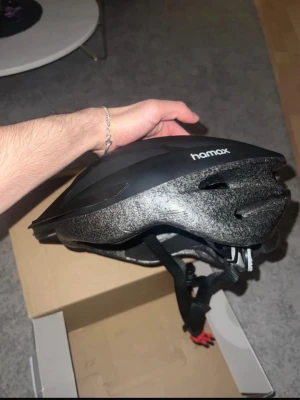 Svart cykelhjälm från Hamax - Snygg svart cykelhjälm från Hamax med ventilerande hål och justerbara remmar. Tillverkad i tålig plast och skum för bra skydd och komfort. Passar perfekt för cykelturer och har en modern, sportig design.