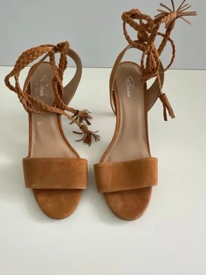 Bruna Rizzo sandaletter i äkta skinn, 39 - Superfina bruna / sandfärgade högklackade skor / sandaler från Rizzo i äkta mocka / skinn / läder. Storlek 39. Superfin flätad snöring runt ankeln med charmig tofs-detalj. Klassiskt tidlöst smala klackar med praktisk diskret förstärkning. Litet märke från envis etikett på ena innersulan, men inget som syns när de är på. Helt NYA!! Nypris 1800:-  Skickas mot porto, men kan även hämtas i Stockholm.