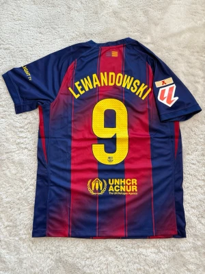 Barcelona matchtröja Lewandowski #9 - Säljer en FC Barcelona matchtröja från Nike med Lewandowski och nummer 9 på ryggen. Tröjan har klassiska blå och röda ränder, klubbmärke och Spotify-logga på bröstet samt UNHCR-logga nedtill. Tillverkad i lätt och ventilerande material, perfekt för fotboll.