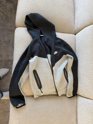 Nike tvåfärgad hoodie med dragkedja - Snygg hoodie från Nike i svart och ljusgrått med dragkedja framtill och två fickor med svarta dragkedjor. Klassisk Nike-logga på bröstet. Tillverkad i mjukt bomullsmaterial och har en stor huva för extra komfort.