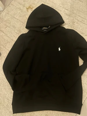 Svart hoodie från Polo Ralph Lauren - Snygg svart hoodie från Polo Ralph Lauren med klassisk vit broderad logga på bröstet. Hoodien har huva och en stor magficka framtill. Perfekt för en stilren och avslappnad look.