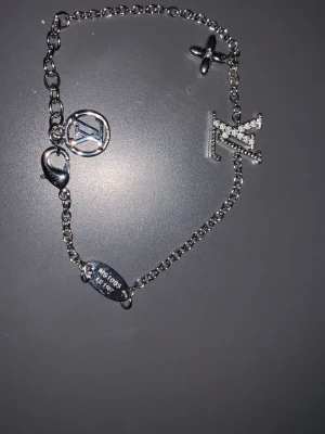  silverfärgat armband - Snyggt armband  silverfärgad metall med kedja och flera hängande berlocker, bland annat LV-loggan och en blomma. Armbandet har glittrande stenar på LV-bokstaven och en rund berlock med ingraverad text. Perfekt för dig som gillar exklusiva accessoarer.