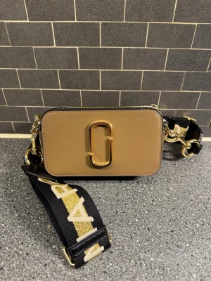 Marc Jacobs Snapshot axelväska beige - Säljer en beige och vit Marc Jacobs Snapshot axelväska i läder med guldiga metalldetaljer och bred svart axelrem med logga i guld. Väskan har två fack med dragkedja och en ficka på baksidan. Perfekt för dig som vill ha en trendig och praktisk väska.
