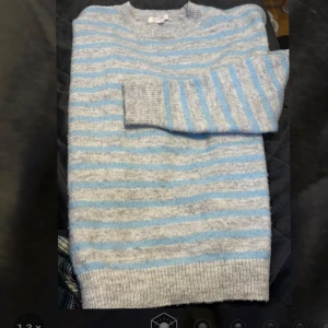 Gråblå randig stickad tröja - Mysig stickad tröja med horisontella ränder i ljusgrått och ljusblått. Tröjan har rund halsringning, långa ärmar och ribbade muddar vid ärmslut och nederkant. Perfekt för lager-på-lager och chill dagar. Tröjan är lite större i storlek.