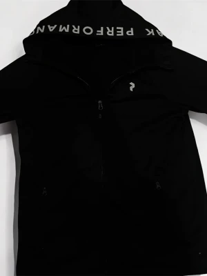 Svart vindjacka från Peak Performance - Svart vindjacka med huva från Peak Performance. Jackan har dragkedja framtill, två sidofickor med dragkedja och vit logga på bröstet. På insidan av huvan står det 'PEAK PERFORMANCE' i vitt. Perfekt för blåsiga dagar.