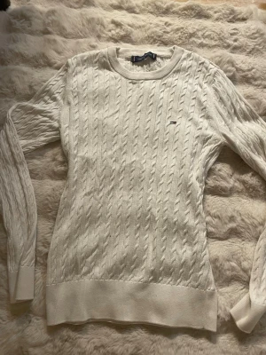 Vit kabelstickad tröja Tommy Hilfiger - . Snygg vit kabelstickad tröja från Tommy Hilfiger med rund halsringning och diskret logga på bröstet. Tröjan har långa ärmar och ribbade muddar vid ärmslut och nederkant. Perfekt för en stilren och klassisk look. Köpt i Turkiet 