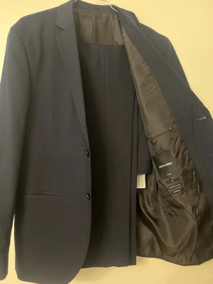 Mörkblå kavaj från jack&jones - Stilren mörkblå kavaj från jack&jones med två knappar framtill, smala slag och innerfoder i svart. Klassisk passform med två fickor fram och en bröstficka. Perfekt för en dressad look.