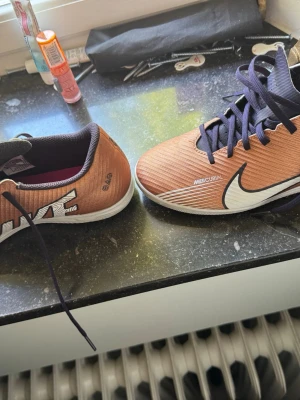 Nike Mercurial bruna fotbollsskor - Säljer ett par Nike Mercurial fotbollsskor i brunt med vita och svarta detaljer. Skorna har snörning och en låg profil, perfekt för snabba rörelser på planen. Tillverkade i syntetmaterial för bra grepp och känsla. Snygg design med stor vit Nike-logga på sidan. Bra för fotboll inomhus. (Pris kan diskuteras)