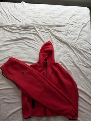 Röd Nike tech set  - Säljer ett rött loungewear set från Nike med matchande hoodie och byxor. Hoodien har dragkedja, huva och svarta detaljer vid ärmslut och fickor. Byxorna har resår i midjan och diskret Nike-logga. Perfekt för chill dagar hemma eller när du vill vara bekväm.