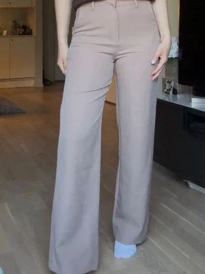 Beige vida kostymbyxor med hög midja - Snygga beige kostymbyxor med hög midja och vida ben. Byxorna har hällor för skärp. Perfekta för en stilren och trendig look. Storlek: S. Längd: 30.