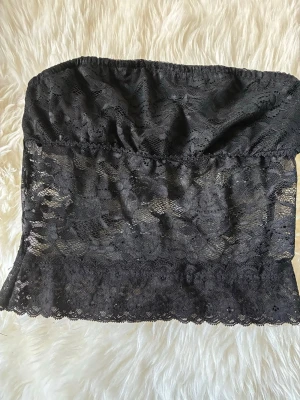 Svart spetsbustier bandeau - Säljer en svart bandeau bustier i genombruten spets med elastisk överdel och vacker blommig spetsdetalj. Modellen är axelbandslös och har en mjuk, stretchig passform som sitter bekvämt mot kroppen. Perfekt att bära under en skjorta eller som den är.