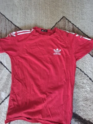 Röd Adidas t-shirt med vita detaljer - Snygg röd t-shirt från Adidas med klassiska vita ränder på axlarna och Adidas-logga på bröstet. T-shirten har rund hals och korta ärmar, perfekt för en sportig och avslappnad stil.