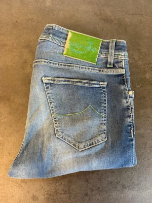 Blåa jeans från Jacob Cohen W35 - Snygga blå jeans från Jacob Cohen med klassisk femficksmodell och gröna detaljer, inklusive en grön patch bak och grön söm på bakfickan. Jeansen har en lätt tvättad look och raka ben. Perfekta för en avslappnad stil. Skriv för mått!