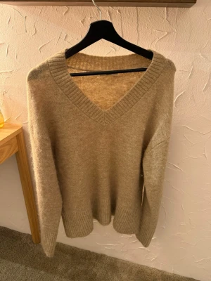 Beige stickad tröja med v-ringning - Mysig beige stickad tröja med djup v-ringning och breda ribbade muddar vid ärmslut och nederkant. Perfekt för lager-på-lager och enkel att matcha med jeans eller kjol. Tröjan har en avslappnad passform och är superbekväm.