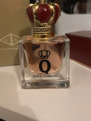 Dolce & Gabbana - Lyxig parfym från Dolce & Gabbana, Q Eau de Parfum. Flaskan är fyrkantig i klart glas med ljusrosa doftvätska och en unik kronformad kork i rött och guld. Förpackningen är elegant röd med gulddetaljer och kronmotiv. Perfekt för dig som gillar exklusiva dofter.