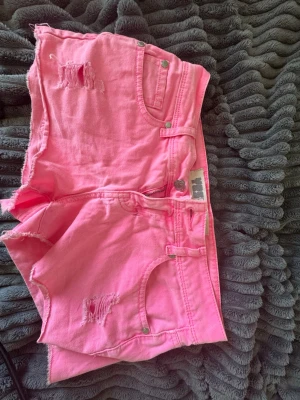Rosa denimshorts med slitningar - Snygga rosa denimshorts med råa kanter och slitningar framtill. Klassisk femficksmodell med bälteshällor och knappgylf. Bakfickorna har dekorativa sömmar. Perfekta för sommaren och ger en cool vibe till din outfit.