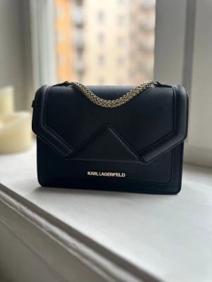 Väska från Karl Lagerfeld - Snygg svart väska från Karl Lagerfeld med guldfärgad kedja och stilren design. Väskan är tillverkad i saffianoläder har geometriska detaljer samt logotyp i guld framtill. Knappt använd så säljes i toppskick och dustbag medföljer. Nypris var ca 3000 kr. 