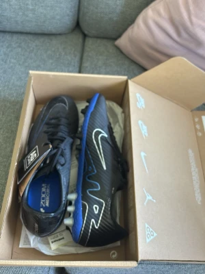 Nike Mercurial svart/blå fotbollsskor - Säljer ett par Nike Mercurial fotbollsskor i svart med blå och vita detaljer. Skorna har en modern design med syntetiskt material och tydlig Nike-logga på sidan. Perfekta för dig som vill ha grymt grepp och stil på planen.
