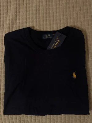 Mörkblå t-shirt från Polo Ralph Lauren - Klassisk mörkblå t-shirt från Polo Ralph Lauren med gul broderad logga på bröstet. T-shirten har rund halsringning och är tillverkad i mjuk bomull för en bekväm känsla. Perfekt för en stilren och avslappnad look.