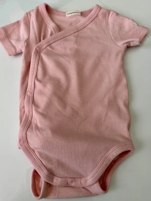 Rosa omlottbody med kort ärm - Söt rosa ribbad omlottbody med korta ärmar och tryckknappar framtill och i grenen. Perfekt för en bekväm och smidig stil. Mjukt material som känns skönt mot huden.