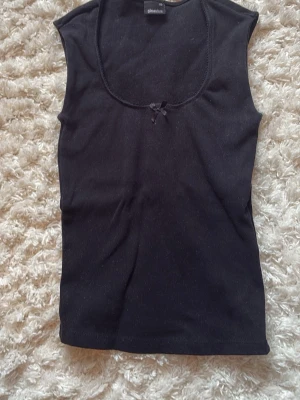 Svart ribbad linnetopp Gina Tricot - Svart ribbad linnetopp från Gina Tricot som ej säljs längre med rundad halsringning och en liten rosett framtill. Endast använd några fåtal gånger, alltså mycket bra skick.