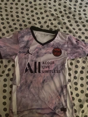 PSG Mbappé matchtröja rosa/lila - Säljer en PSG matchtröja med Mbappé och nummer 7 på ryggen. Tröjan har ett unikt rosa och lila marmormönster, svart krage och detaljer. Officiella loggor från PSG, Jordan och sponsortryck på bröstet. Perfekt för dig som älskar fotboll och vill sticka ut på planen. Priset går att snacka om 