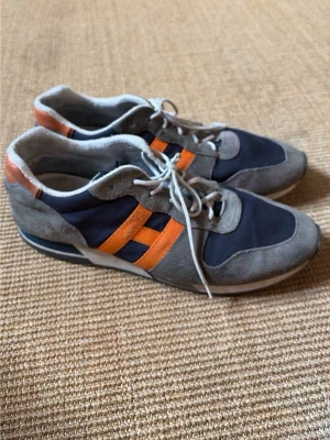 Grå och marinblå sneakers med orange detaljer - Snygga Hogan sneakers i grå mocka och marinblå textil med orangea detaljer på sidorna. Skorna har vita snören och låg profil, perfekt för en sportig och avslappnad stil. Märket syns som ett H på sidan. Riktigt fräscha färgkombos för dig som gillar streetwear. Nypris 7000