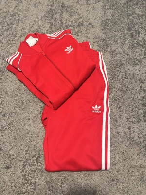 Röd Adidas tracksuit med vita ränder - Säljer en klassisk röd Adidas tracksuit med vita ränder längs ärmar och ben. Setet består av en zip-jacka och matchande byxor, båda med Adidas logga och retrodesign. Perfekt för chill dagar hemma eller streetwear. Materialet är mjukt och stretchigt för maximal komfort.