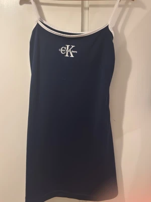 Marinblå klänning från Calvin Klein - Snygg marinblå klänning från Calvin Klein med vita tunna axelband och vit kant upptill. Klänningen har en enkel, rak passform och är dekorerad med CK-logga på bröstet. Perfekt för en stilren look.