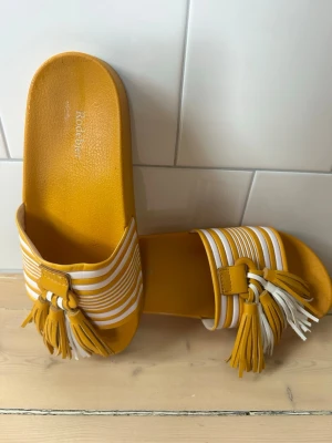 Randiga sandaler med tofsar från Rodebjer - Ikonisk sandal från Rodebjer i den härliga gula färgen. Sparsamt använda med helt intakta tofsar och inga märken av fötter i fotbäddarna. Tecken på användning enbart på sulan undertill. 