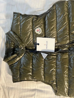 Moncler väst - Snygg olivgrön dunväst från Moncler med glansig finish och klassisk logotyp på bröstet. Västen har två dragkedjeförsedda fickor framtill och hög krage. Perfekt för lager-på-lager och streetstyle.