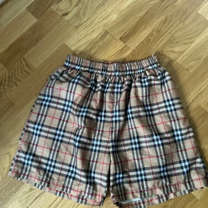 Burberry badshorts - Snygga burberry shorts med klassiskt rutigt mönster i svart, vitt och rött. De har en bekväm resår i midjan och är tillverkade i ett lätt och svalt material, perfekt för varma dagar. Passformen är normal och stilen är tidlös med en touch av preppy vibe.