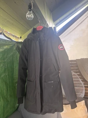 Svart parkas från Canada Goose - Svart parkas från Canada Goose med huva och två stora fickor framtill. Klassisk logotyp på vänster ärm. Lång modell med dragkedja och knappar, samt ribbade muddar vid ärmslut. Perfekt för kyliga dagar och stilren look.