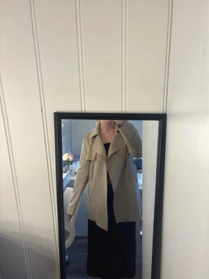 Beige trenchcoat med bälte - Stilren beige trenchcoat med klassisk krage och bälte i midjan. Jackan har två bröstfickor med lock och en rak passform som ger en snygg siluett. Perfekt för dig som gillar en enkel men trendig look. Materialet ser ut att vara ett mjukt tyg, troligen polyester eller bomullsblandning.